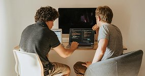 Two people sitting side by side at a desk, working on laptops displaying code, with a large monitor in the background. Both are focused on their work in a well-lit room.