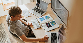 A person sits at a desk using a computer mouse, with two laptops, a keyboard, and an open notebook displaying maps and charts in front of them, suggesting they are studying or working.