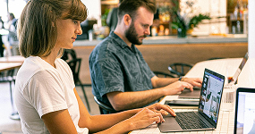 Webtrix Innovations Two people sit side by side at a table, working on their laptops in a bright, modern indoor setting. The woman in front is typing, while the man behind her focuses on his screen.