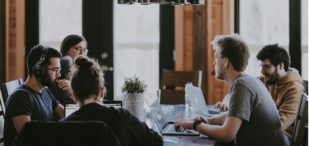 Webtrix Innovations Five people sit around a table working on laptops, some wearing headphones. There is a small plant in the center of the table, and the room has large windows letting in natural light.